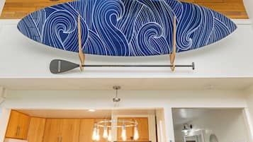 Un adorno mural con forma de tabla de surf, un remo, una cocina moderna con armarios de madera y una lámpara colgante.