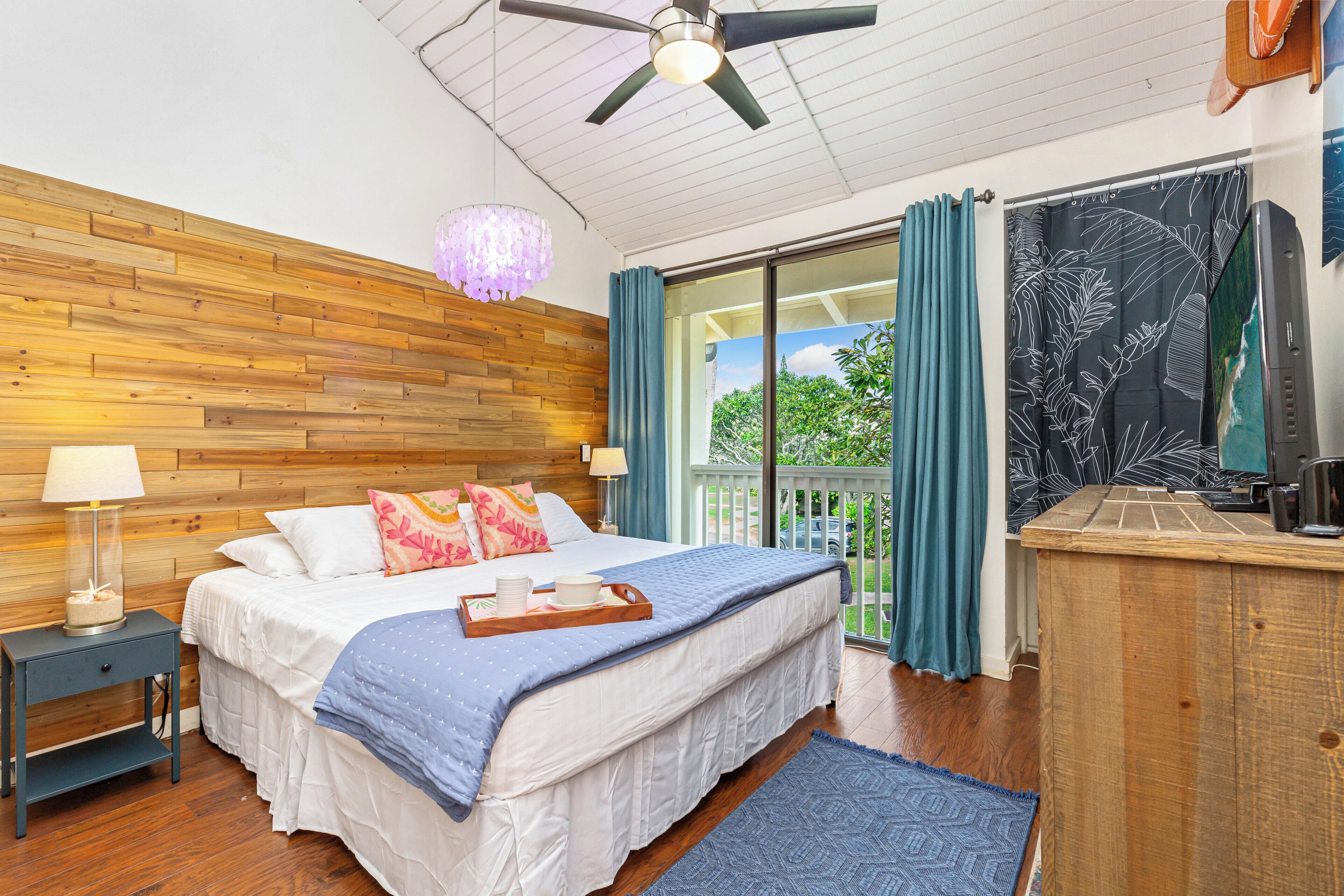 A bedroom with a wooden headboard, a bed with white bedding and blue accents, a nightstand with a lamp, a dresser, and a view of greenery through the window.