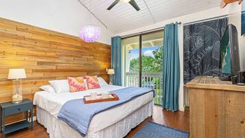 A bedroom with a wooden headboard, a bed with white bedding and blue accents, a nightstand with a lamp, a dresser, and a view of greenery through the window.