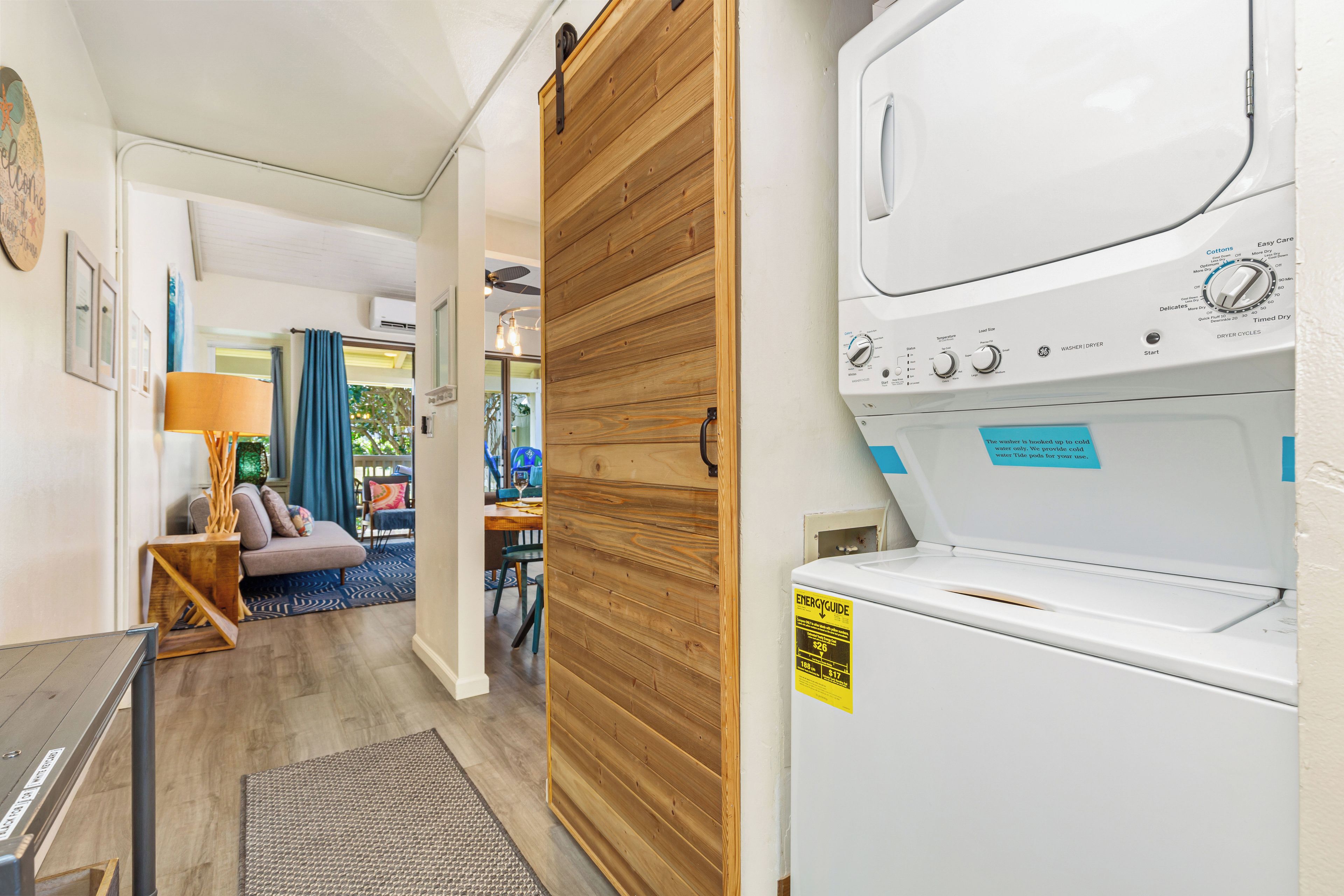 Un área de lavandería con lavadora y secadora, una puerta de madera y vistas a una sala de estar con sofá y lámpara.