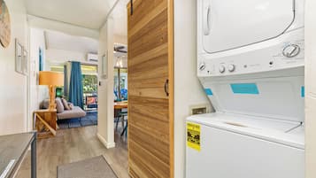 Un área de lavandería con lavadora y secadora, una puerta de madera y vistas a una sala de estar con sofá y lámpara.