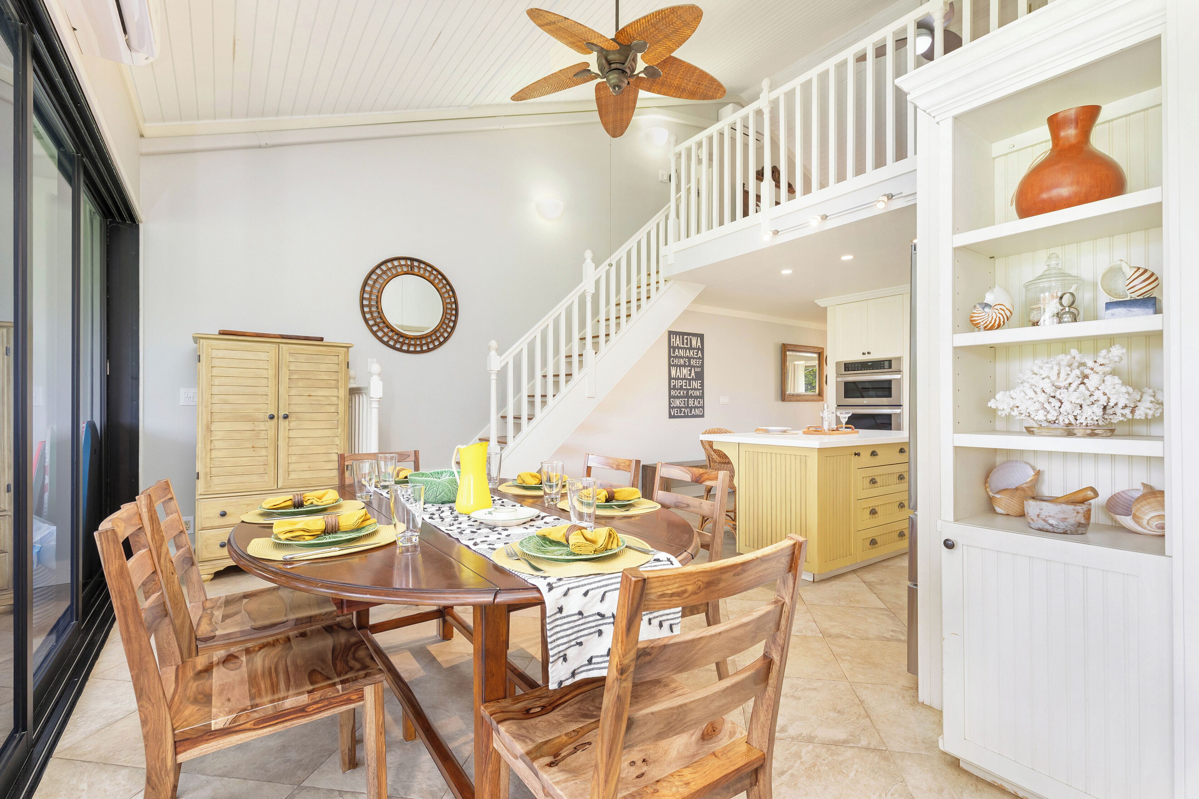 A dining area with a wooden table set for six, surrounded by wooden chairs. A staircase with white railings leads to an upper level.