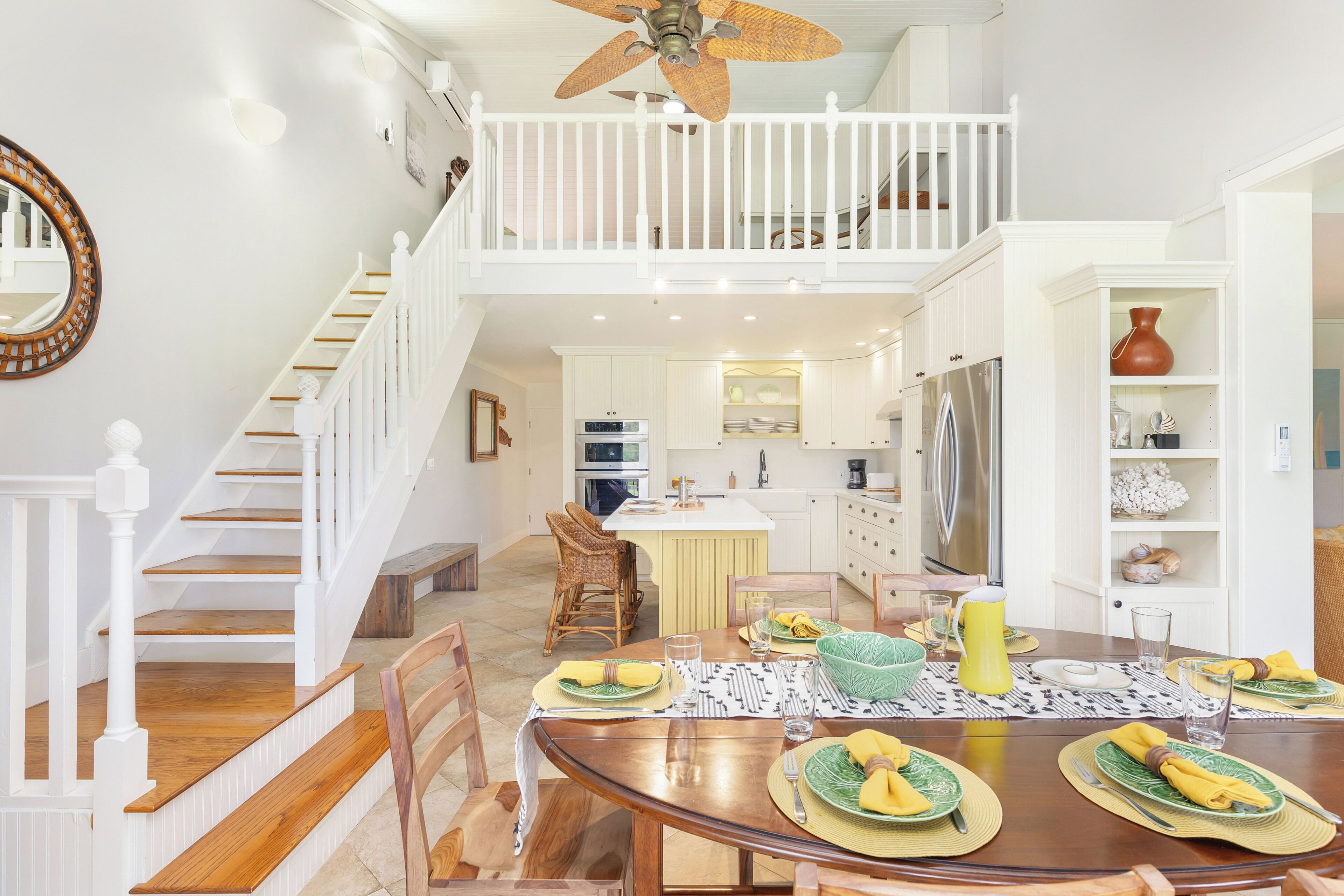 Une vaste salle à manger dotée d’un haut ventilateur de plafond, d’un escalier menant à un niveau supérieur et d’une cuisine à l’arrière-plan.