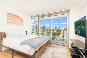 A modern bedroom with a large bed, a city view through floor-to-ceiling windows, a flat-screen TV, and a painting on the wall. - Chic Studio With Skyline Views at The Park on Keeaumoku (Honolulu)