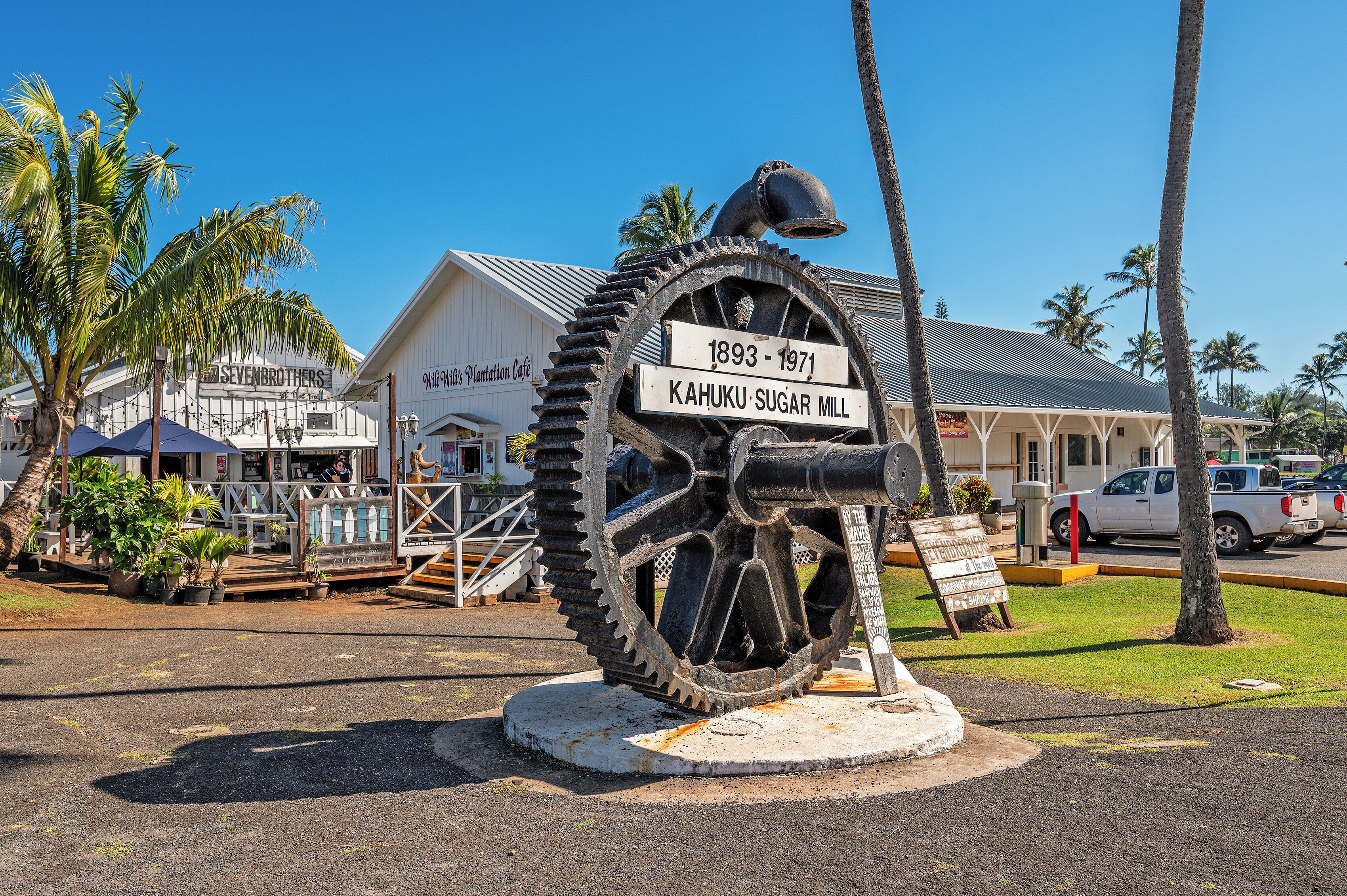 A historical marker for the Kahuku Sugar Mill, a white building with a signboard, and a white pickup truck parked nearby.