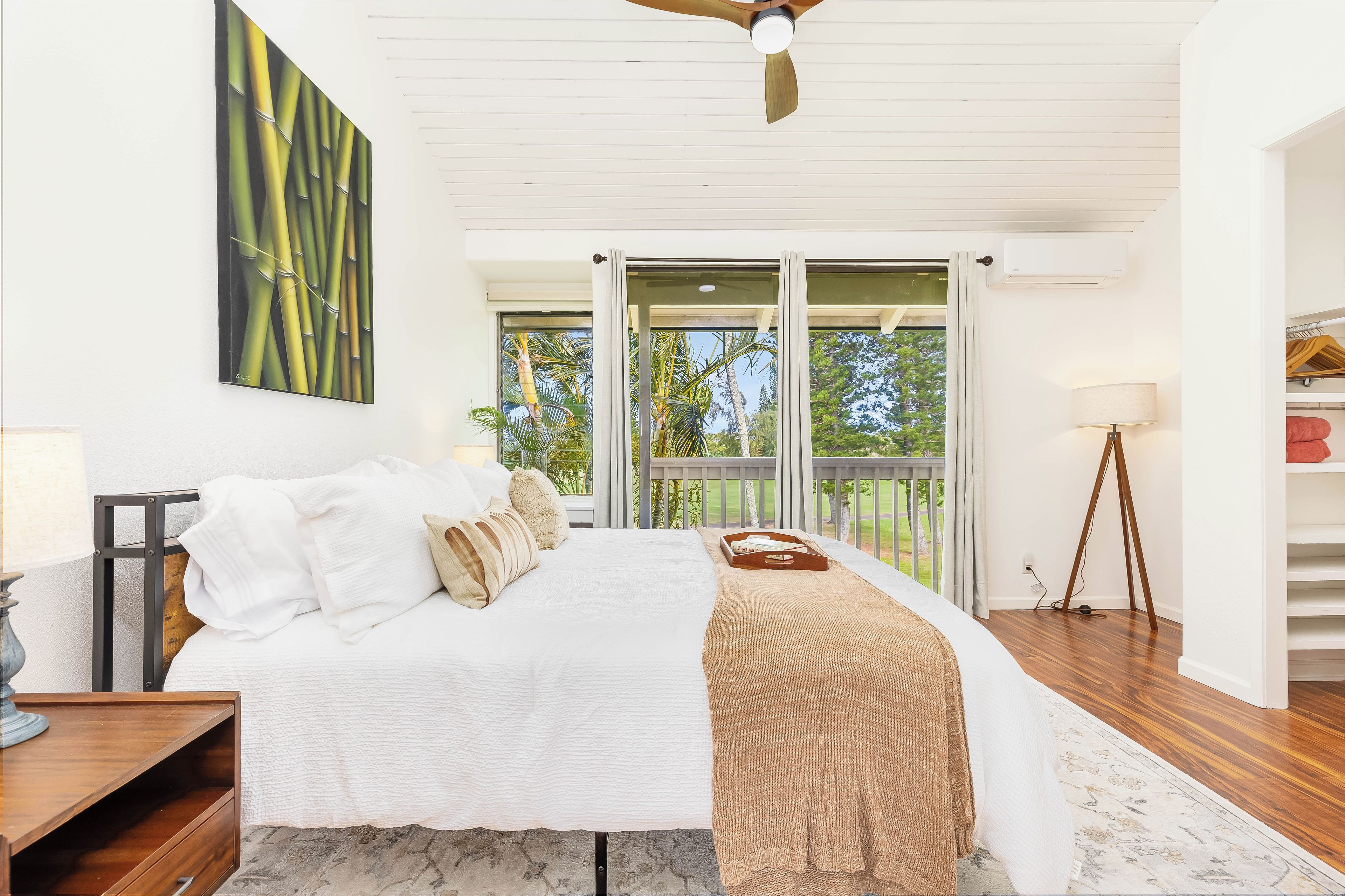 Une chambre à coucher comprenant un lit, une table de chevet, une lampe, un ventilateur de plafond et une grande fenêtre donnant sur un paysage verdoyant.