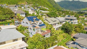A residential area with houses featuring solar panels, surrounded by lush greenery and hills.