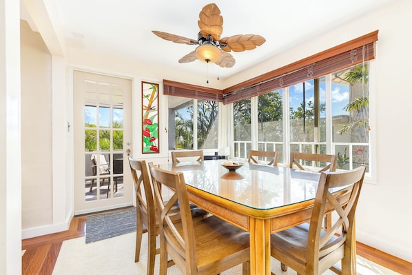 A dining room with a glass table, wooden chairs, a ceiling fan, and large windows with a view of greenery.