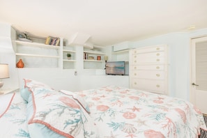 A bedroom with a bed, a dresser, a TV, and shelves with books and decorative items.