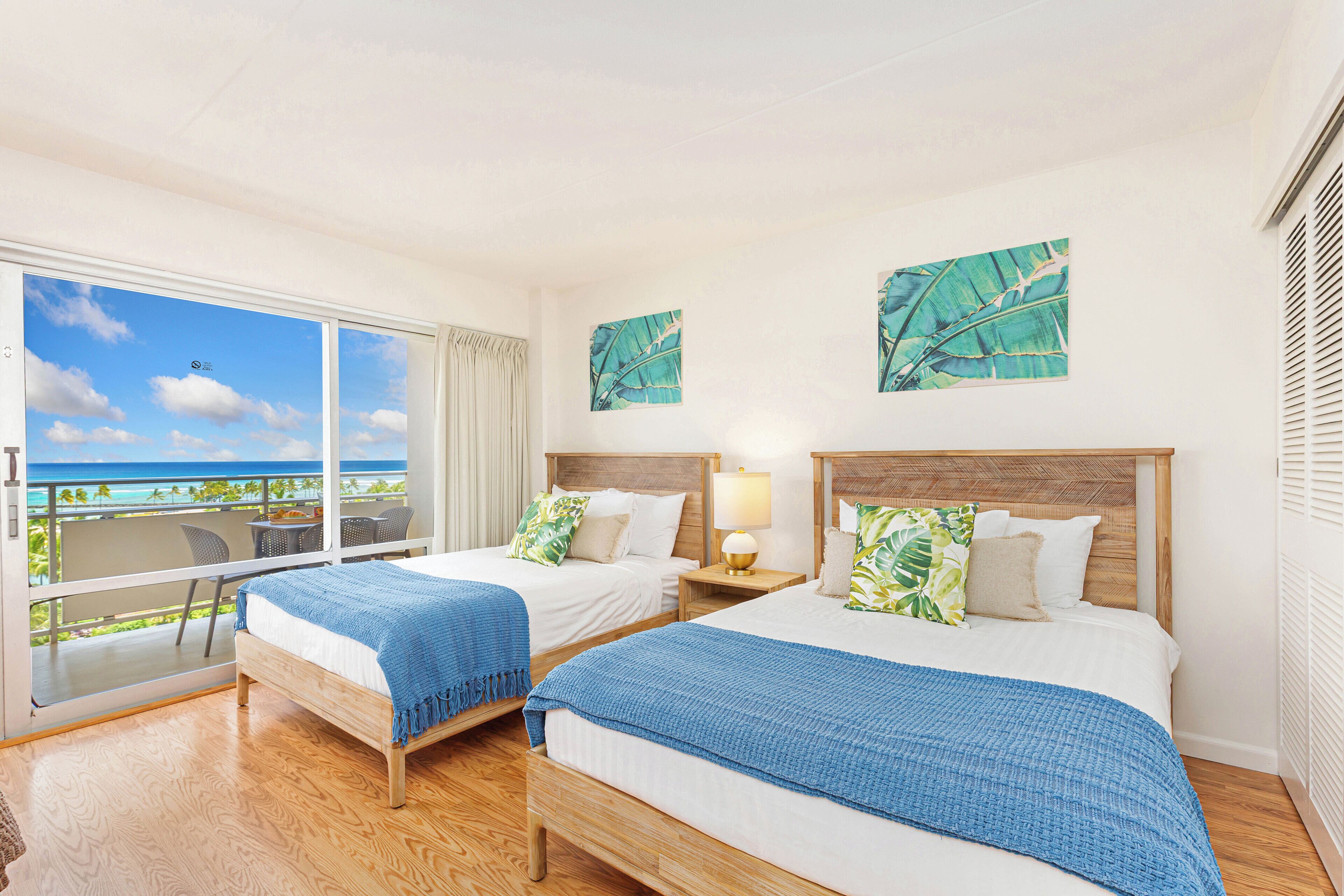 A hotel room with two beds, a balcony with a view of the ocean, and tropical-themed wall art.