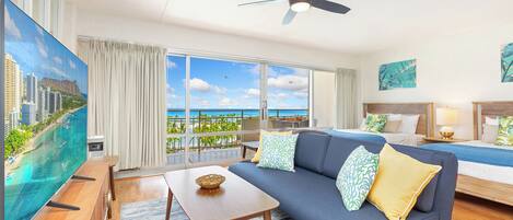 A modern hotel room with a large TV, a sofa, a bed, and a balcony with a view of the ocean.