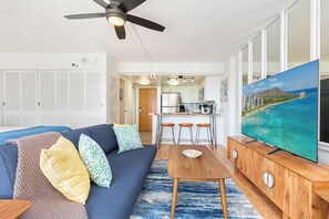 A modern living room with a sofa, a coffee table, and a TV on a wooden cabinet. There is a kitchen area with a bar and stools. - Skyline-lagoon and Fireworks Magic From the Ilikai W/top-tier Amenities (Honolulu)