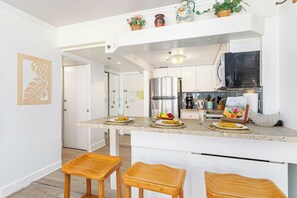 A modern kitchen with white cabinetry, a stainless steel refrigerator, and a granite countertop. There are two wooden stools and a small island with a fruit bowl and a coffee maker. - Stunning Ocean & Fireworks Lagoon View From Condo at Ilikai W/full Kitchen (Honolulu)