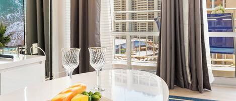A hotel room with a dining table, two wine glasses, a baguette, cheese, and a cutting board.
