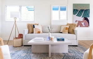 A living room with a light-colored sofa, a coffee table with books and a vase, a floor lamp, and a large window with blinds. - Serene Lanikai House Near the Beach W/outdoor BBQ & Close to Shopping Center (Kailua)