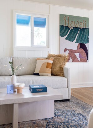 A living room with a sofa, a coffee table with a vase and a book, a window with a view, and a wall with a Hawaii artwork. - Serene Lanikai House Near the Beach W/outdoor BBQ & Close to Shopping Center (Kailua)