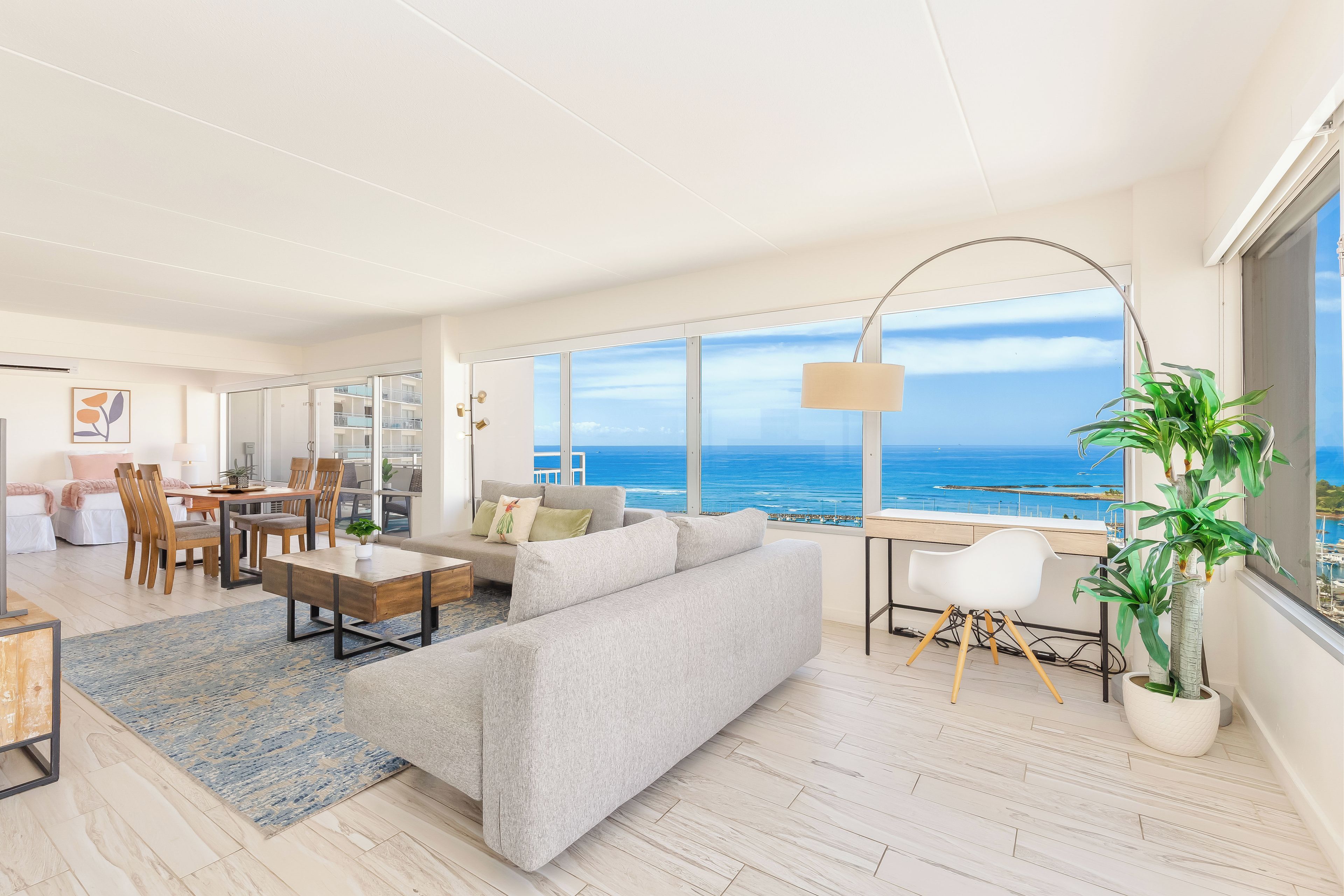 A spacious living area with a large window overlooking the ocean, a sofa, a dining table, and a potted plant.