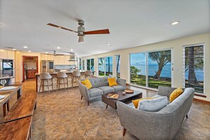 A spacious living area with a grey sofa set, a wooden coffee table, and a dining table with benches. There is a ceiling fan and a large window with a view of the sea. - Oceanfront Gem in the Beautiful Kaneohe Dream Home Paradise Family Retreat (Kaneohe)