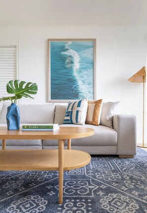 Sala de estar com sofá, mesa de centro, quadro da paisagem marítima, planta em vaso e luminária de chão.