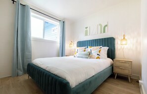 Cama bem arrumada com cabeceira azul-turquesa, roupa de cama branca e almofadas decorativas. Há duas luminárias de mesa com design trançado. O quarto tem paredes brancas, uma janela com cortina e piso de madeira.