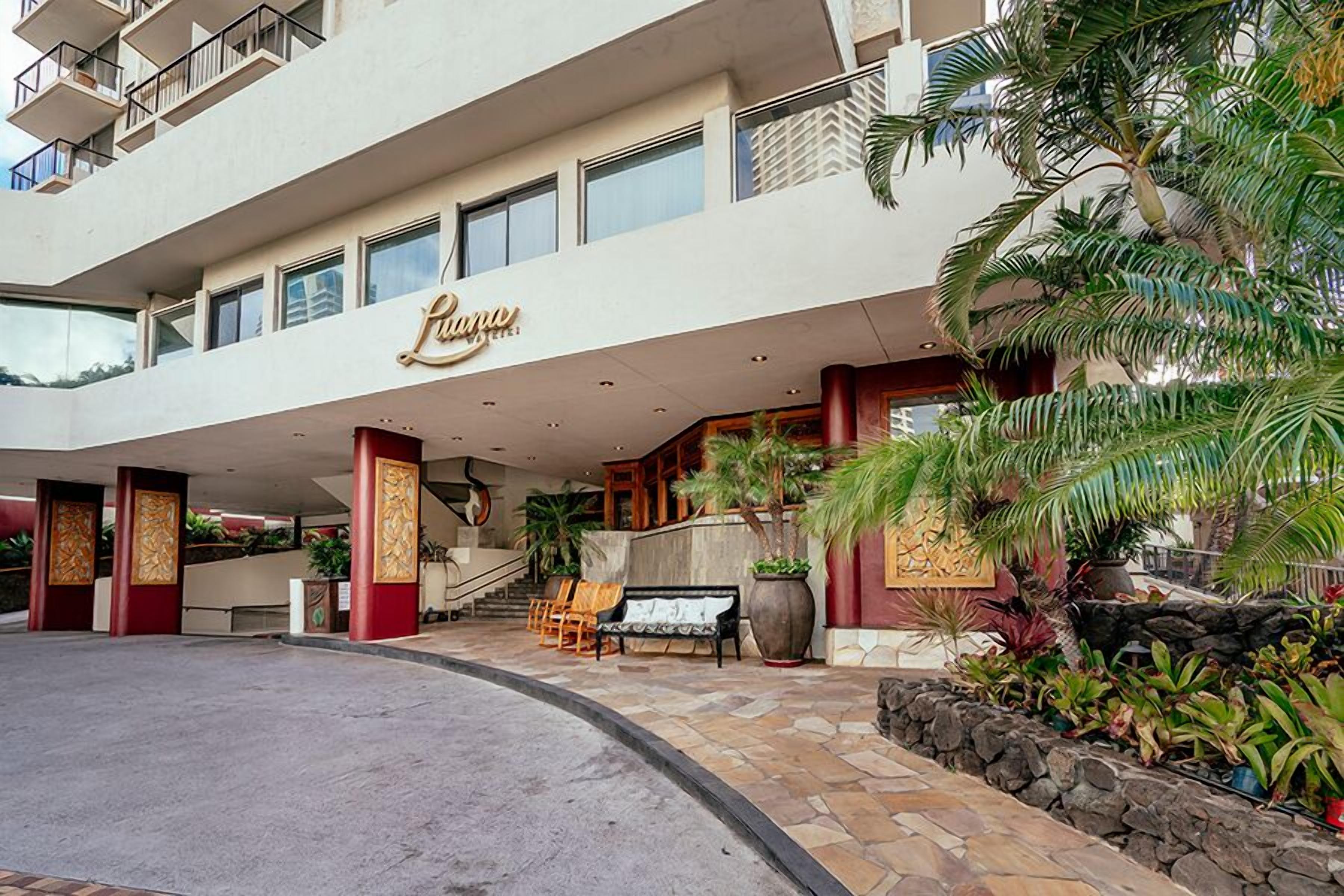 Entrada de hotel con una fachada imponente, una palmera y un banco.