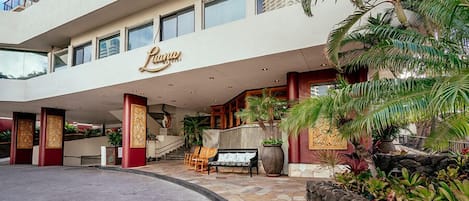 Entrada de hotel con una fachada imponente, una palmera y un banco.