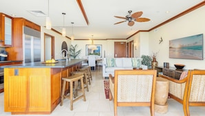 A spacious kitchen with a central island, wooden bar stools, a dining area with a white sofa, and a ceiling fan.