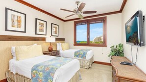 A hotel room with two beds, a ceiling fan, a TV, a window with a view, and framed pictures on the wall.