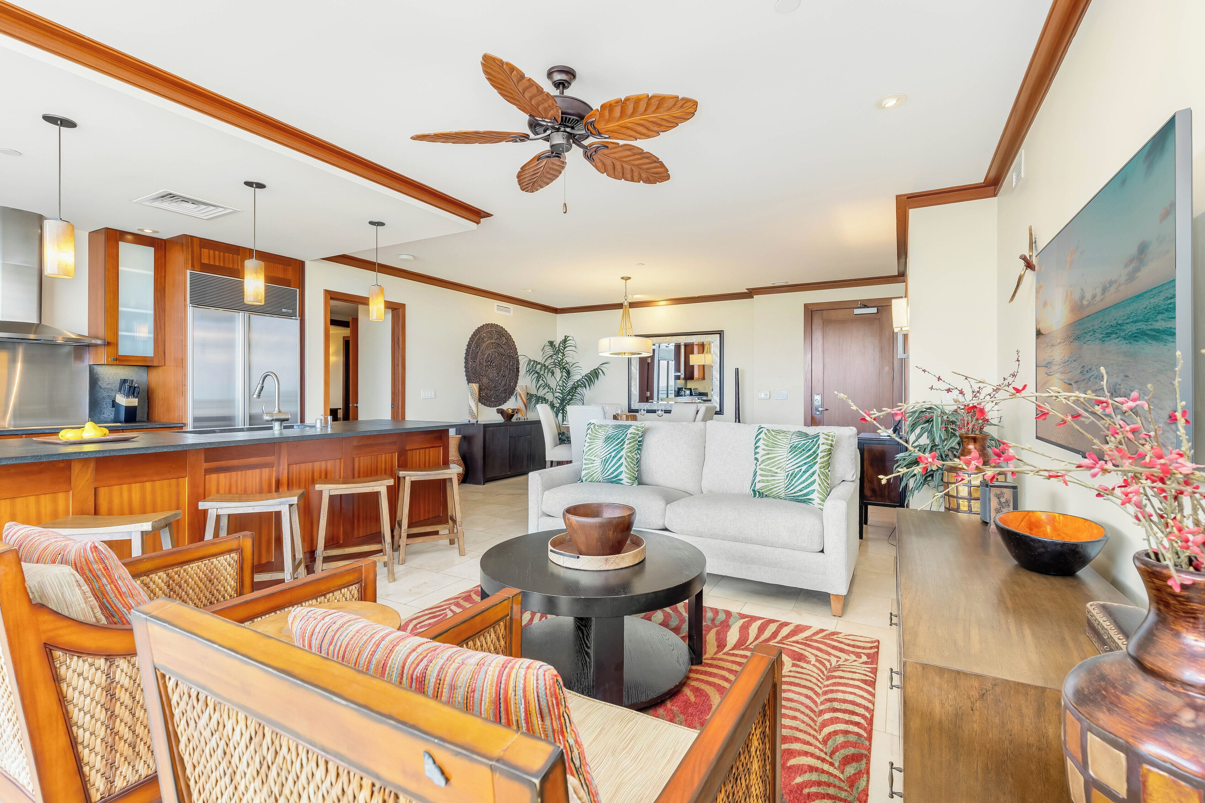 A spacious living area with a central seating arrangement, a kitchen in the background, and a ceiling fan.