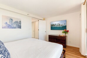 A bedroom with a large bed, a wooden dresser, a painting of a beach, and a door leading to another room. - Diamond Head & Golf Course Views at Island Colony W/amazing Amenities (Honolulu)