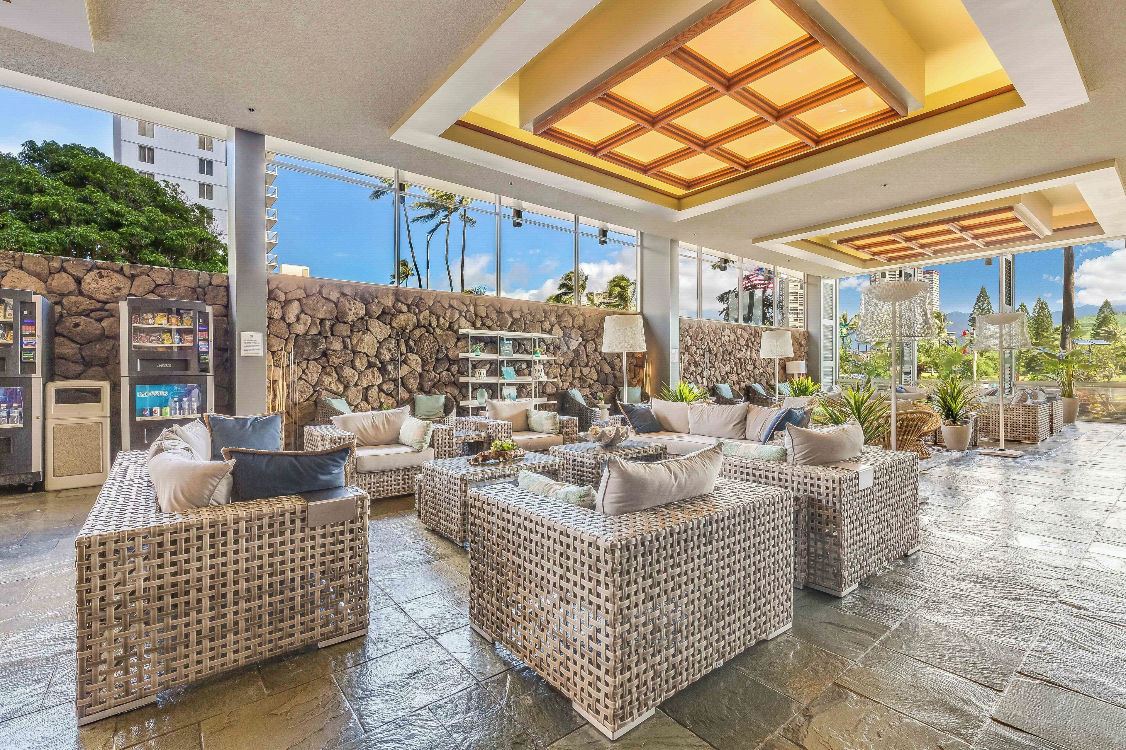 A spacious lounge area with wicker furniture, a stone wall, and a view of palm trees and buildings.