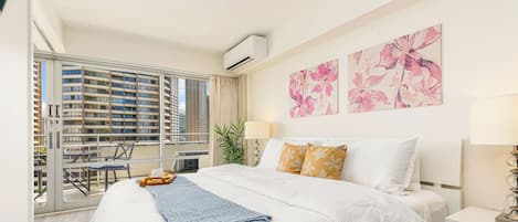 A modern bedroom with a large bed, a bedside table, a window with a view of high-rise buildings, and a balcony with outdoor furniture.