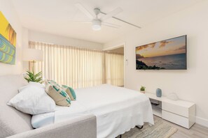 A modern bedroom with a bed, a ceiling fan, a bedside table, a nightstand, a lamp, a painting, and a view of the ocean. - Renovated City Suite at Ilikai Steps to the Beach & Short Walk to Restaurants (Honolulu)