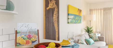 A modern living room with a wooden dining table, a fruit bowl, a framed picture, and a sofa with cushions.