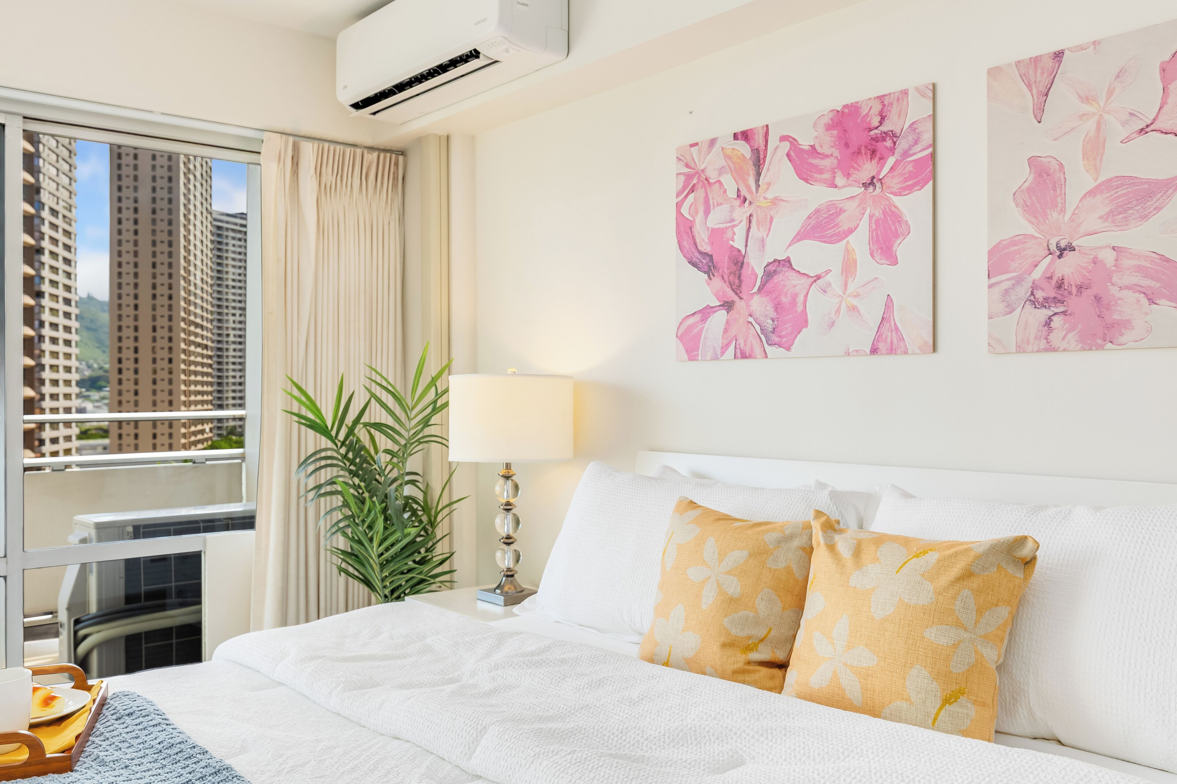 A hotel room with a bed, a window with a view of tall buildings, a plant, and floral artwork on the wall.