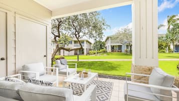 A patio with white furniture, a patterned rug, and a view of a well-maintained lawn and houses in the background.