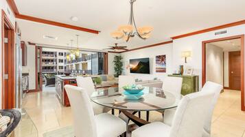 A modern living room with a glass dining table, white upholstered chairs, a chandelier, and a view of the kitchen and balcony.