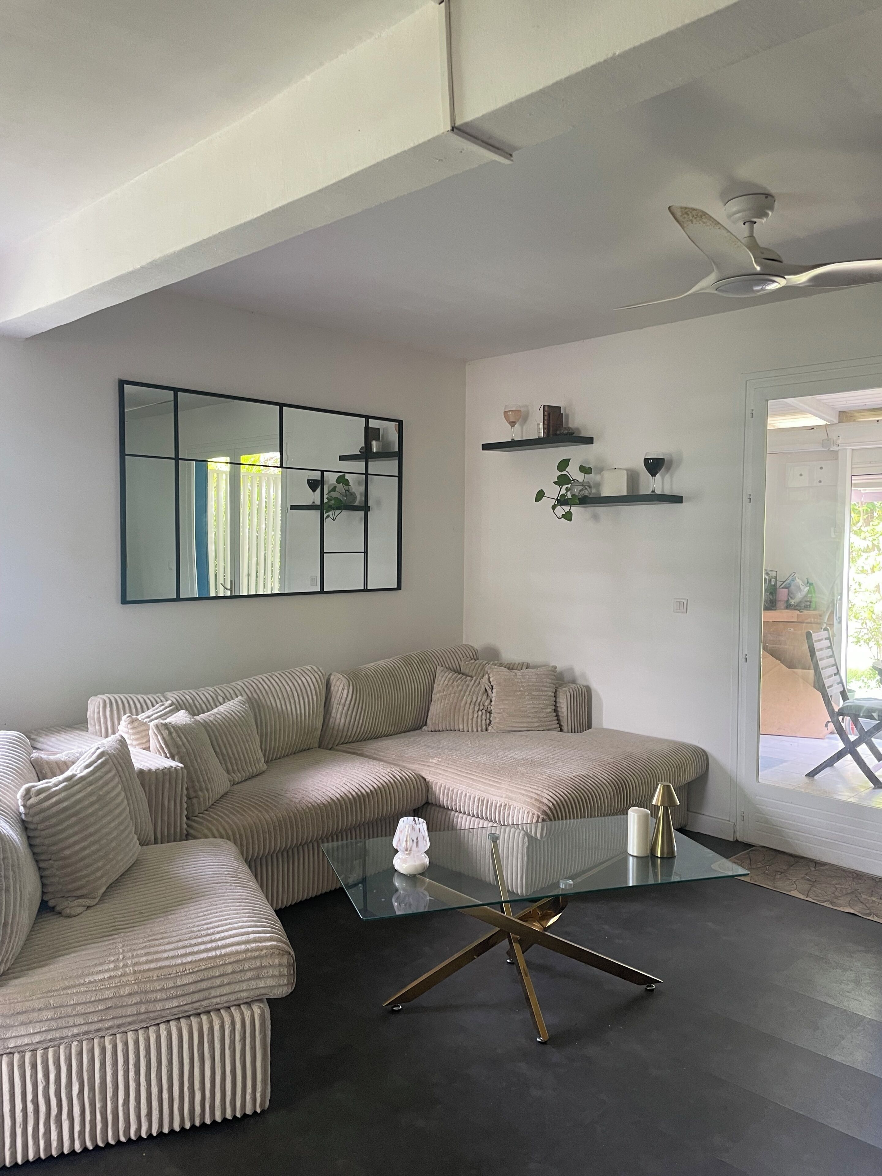 Un salon moderne avec un canapé d’angle, une table basse en verre avec un pied doré et un ventilateur de plafond.