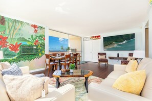 A living room with a large tropical mural, a dining area with wooden furniture, and a large ocean view painting. - Ocean View Condo at Foster Tower Across From Beach W/great Amenities (Honolulu)