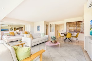 A modern living room with a sofa, a dining table with chairs, a coffee table, and a kitchen area visible through an open door. - Beachfront Property W/sand Access at Waikiki Shore Condo With Full Kitchen (Honolulu)