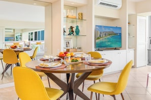 Uma sala de jantar com uma mesa redonda, cadeiras amarelas e uma mesa de vidro. Há uma televisão na parede e uma área de cozinha ao fundo.