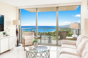 A modern living room with a glass balustrade, a sofa, a coffee table, and a view of the sea and cityscape. - Ocean-sunset Views Designer Condo at The Watermark W/great Building Amenities (Honolulu)