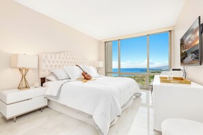 A modern bedroom with a large bed, a guitar, a desk, and a view of the ocean. - Ocean-sunset Views Designer Condo at The Watermark W/great Building Amenities (Honolulu)