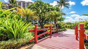 A red wooden bridge leading to a building with multiple balconies, surrounded by lush greenery and palm trees. - Beach Villas Peekaboo Ocean & Mountain View Resort W/luxurious Amenities (Kapolei)