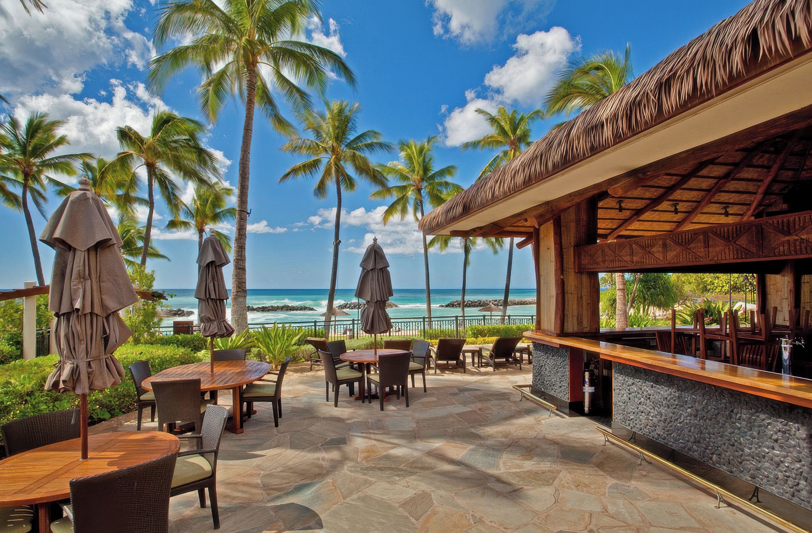 An outdoor patio with tables and chairs, a bar area, and a view of the ocean and palm trees.