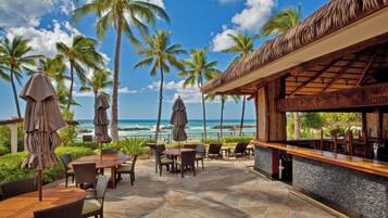 An outdoor patio with tables and chairs, a bar area, and a view of the ocean and palm trees.