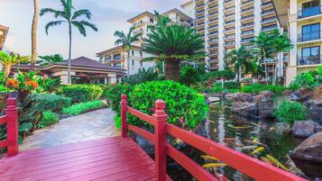A red wooden bridge over a pond with fish, surrounded by lush greenery and palm trees, leading to a multi-story building with balconies.