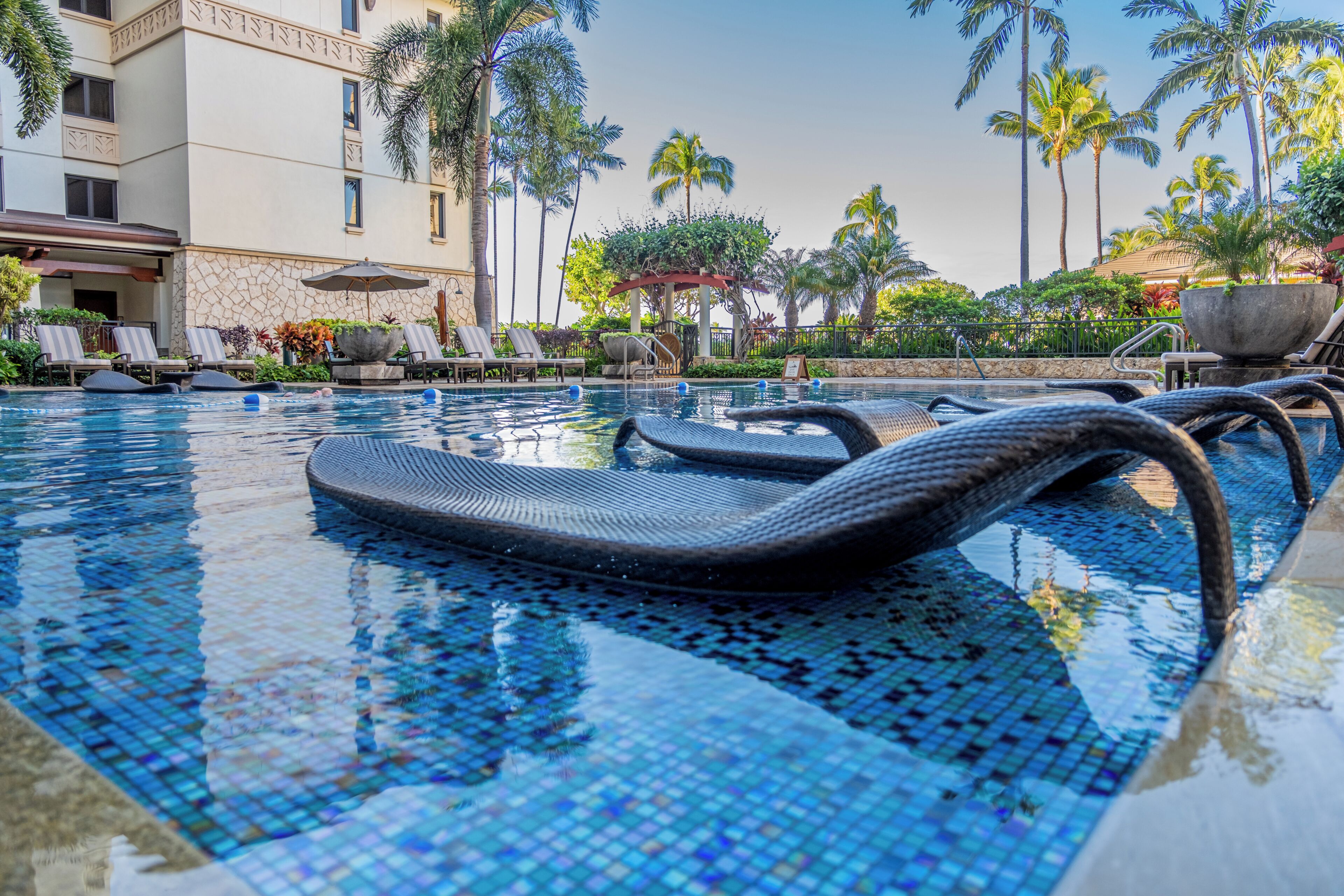 A pool with mosaic tiles, lounge chairs, palm trees, and a building with a stone facade.