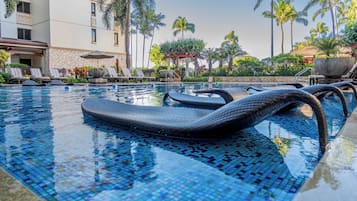 A pool with mosaic tiles, lounge chairs, palm trees, and a building with a stone facade.