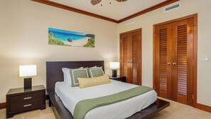 A neatly arranged bedroom with a bed, two bedside tables with lamps, a painting of a beach scene, and wooden doors. - Beach Villas Peekaboo Ocean & Mountain View Resort W/luxurious Amenities (Kapolei)